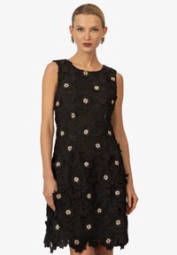 Schwarzes ärmelloses Kleid mit einem strukturierten floralen Spitzenmuster, verziert mit kleinen weißen Blumenakzenten und einem gewellten Saum.