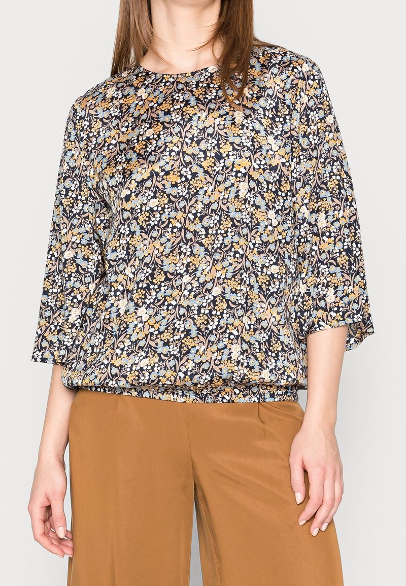 Blusa floreale in navy con motivi gialli, blu e bianchi. Caratterizzata da un colletto rotondo, maniche ¾ e una vestibilità ampia. Il tessuto appare leggero.