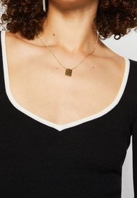 Haut tricot noir avec un col en V et des accents blancs. Un collier en or avec un pendentif rectangulaire se trouve juste en dessous de l'encolure.
