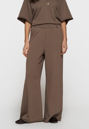 Personne portant un pantalon ample à jambes larges marron et un haut assorti à manches courtes, debout devant un fond uni.