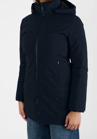 Femme portant une veste d'hiver matelassée bleu marine avec une fermeture éclair à l'avant et une capuche, associée à un jean en denim bleu, se tenant devant un fond blanc.