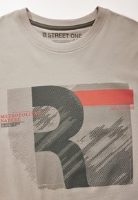 Ljusgrå bomullströja med ett grafiskt mönster i mörkgrått och rött. Texten inkluderar "METROPOLITAN NATURE" och ett stort bokstav "R" i en strukturerad stil.