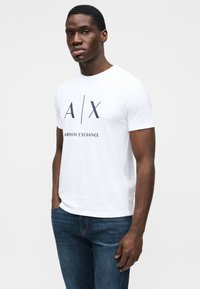 Tricou alb din bumbac cu mâneci scurte, având imprimat pe față un logo „A/X” albastru închis și textul „ARMANI EXCHANGE”. Asortat cu blugi albaștri.