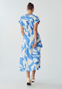Bijela haljina s plavim apstraktnim uzorcima lišća, kratkim rukavima, nabranim strukom i slojevitim rubom, uparena s bijelim potpeticama.