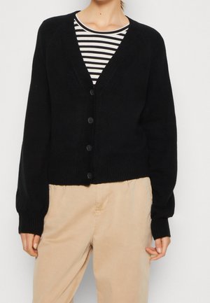 Person trägt eine schwarze, geknöpfte Strickjacke über einem schwarz-weiß gestreiften Oberteil und beige Hosen, dargestellt vom Hals bis zur Hüfte.