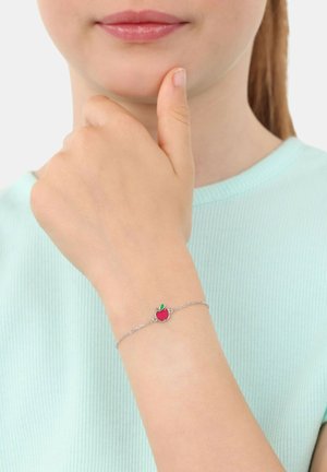 Braccialetto in argento con un charm a forma di mela rossa con una foglia verde, indossato su una mano appoggiata vicino al mento. La parte superiore è di un verde menta.