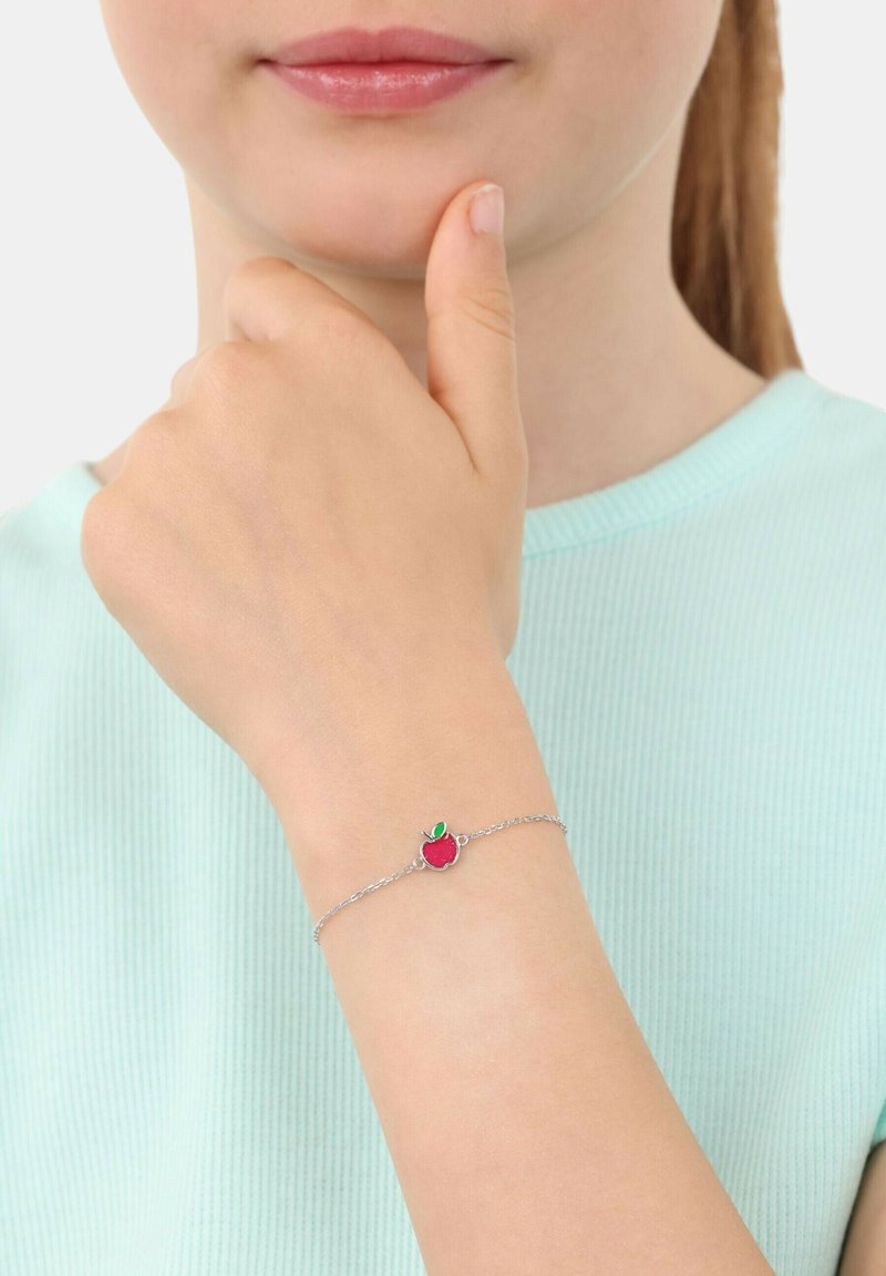 Silver kedja armband med en röd äpple charm med ett grönt blad, buren på en hand som är placerad nära hakan. Toppen är mintgrön.