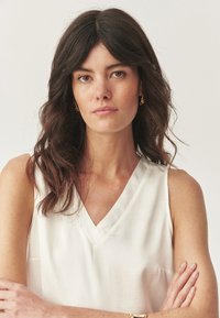 Blouse blanche à col en V avec une texture lisse, portée par un mannequin aux cheveux sombres et ondulés ; bras croisés, design minimaliste et élancé.
