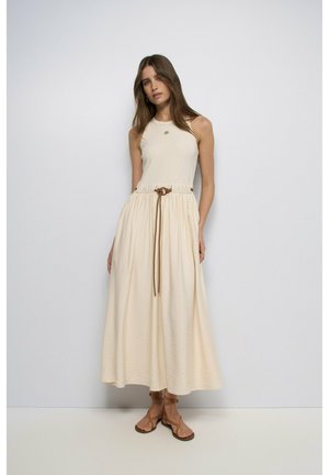 Mujer con top beige sin mangas y falda larga crema con cinturón marrón, de pie frente a un fondo liso gris claro.
