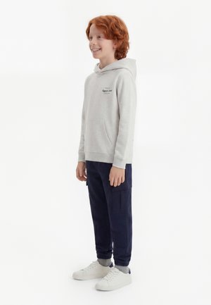 Garçon roux souriant debout portant un sweat à capuche gris clair, un pantalon cargo bleu foncé, des chaussettes grises et des baskets blanches sur fond blanc.