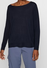 Pull bleu marine avec un col large, texture côtelée sur les manches et coupe décontractée. Main partiellement visible sur un pantalon bleu clair.