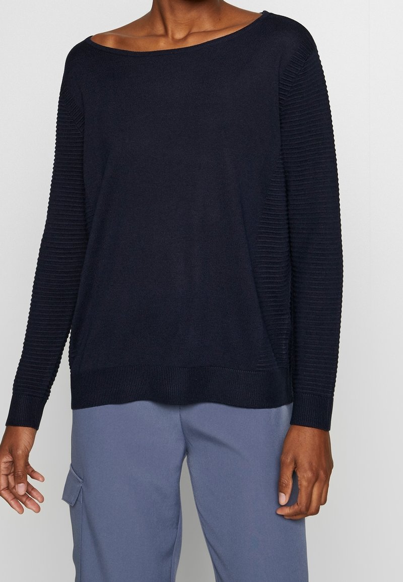Pull bleu marine avec un col large, texture côtelée sur les manches et coupe décontractée. Main partiellement visible sur un pantalon bleu clair.