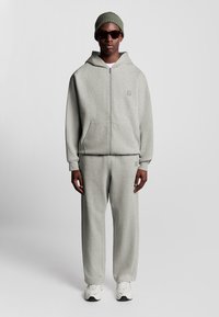 Mand iført en lysegrå lynlås hættejakke og matchende svedbukser, olivengrøn beanie, mørke solbriller og hvide sneakers, stående mod en ensfarvet baggrund.
