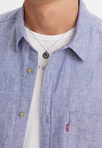 Jasnoniebieska koszula zapinana na guziki z kieszonką, wykonana z teksturowanej tkaniny. Noszona na białej koszulce, z srebrnymi naszyjnikami z zawieszkami.