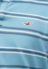 Tissu bleu clair avec des rayures horizontales blanches et bleu marine et un petit logo oiseau rouge brodé sur la zone de la poitrine.
