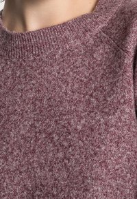 Maroon sweater dengan kerah bulat bergaris, kain bertekstur, dan permukaan yang sedikit berbulu, menyoroti perpaduan warna dalam rajutan.