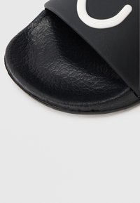 Șlapi negri cu talpă texturată și o curea lată care prezintă un logo alb. Materialul pare sintetic, cu finisaj neted.