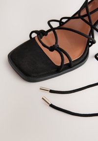 Sandalo in suede nero con punta quadrata e design incrociato con lacci. Caratterizzato da terminali dorati sui lacci e soletta interna marrone a contrasto.