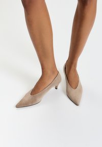 Escarpins en daim beige avec un bout pointu et une découpe en V sur les côtés. Les talons sont courts et fins. Une texture subtile est visible.