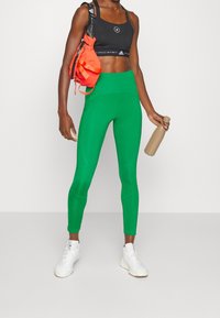 Atletisk person i gröna leggings, svart sportbh, vita sneakers som håller en beige vattenflaska med en orange axelväska.