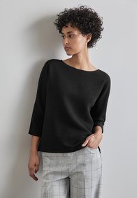 Zwarte textuur top met driekwart mouwen, gecombineerd met lichtgrijze geruite broek, waarmee een casual, modern ontwerp wordt benadrukt.