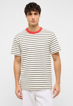 Young man with short curly hair wearing a white t-shirt with black horizontal stripes and a red collar, standing against a plain background.