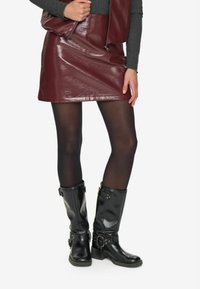 Donna che indossa una gonna di pelle bordeaux, collant neri, stivali di pelle neri a metà polpaccio e un top a maniche lunghe grigio scuro con un gilet coordinato.