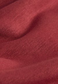 Close-up van zachte, gestructureerde stof in gedempt rood met zichtbaar breipatroon en zachte plooien die schaduwen en diepte creëren.