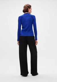 Suéter azul ajustado com um decote alto e textura canelada, combinado com calças largas pretas, visto de costas, exibindo o design.