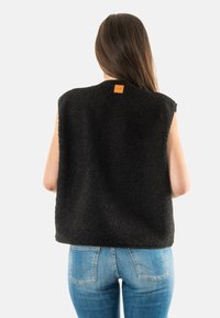 Chaleco de forro polar negro con una superficie texturizada, diseño sin mangas y una pequeña etiqueta de cuero en la parte trasera. Combinado con jeans azules.
