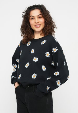 Žena s kučeravými vlasmi, oblečená v čiernej mikine zdobenej bielymi a žltými vzormi sedmokrásky, ruky v vreckách, mierne sa usmieva.