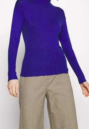 Kvinde iført en tætsiddende lilla ribstrikket polotrøje og beige ternede bukser med brede ben, stående mod en ensfarvet hvid baggrund.