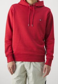 Sudadera de algodón roja con un bolsillo canguro en el frente, capucha con cordón y logo bordado. Textura suave, puños y dobladillo acanalados.