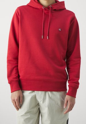 Hoodie en coton rouge avec une poche kangourou à l'avant, capuche à cordon de serrage et logo brodé. Texture lisse, poignets et ourlet côtelés.