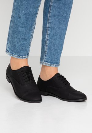 Persoon draagt zwarte leren veterschoenen Oxford met lage hakken en blauwe spijkerbroek tegen een effen lichte achtergrond.