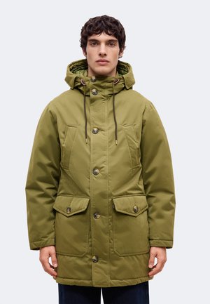 Un homme pose avec une longue parka à capuche vert olive, dotée de cordons de serrage, de gros boutons et de poches à rabat, sur un fond clair et uni.