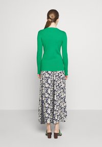 Vihreä istuva neulepaita, jossa pitkät hihat, yhdistetty laivastonsiniseen kukkakuosiseen maxihameeseen, jossa on kermaan vivahtavia kukkia. Asu viimeistelty korollisilla sandaaleilla.