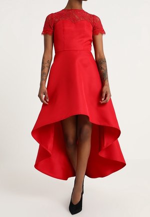 Kobieta ubrana w czerwoną sukienkę z krótszym przodem i dłuższym tyłem z koronkowymi krótkimi rękawami, stojąca w czarnych szpilkach, z tatuażami na ramionach, na jednolitym tle.