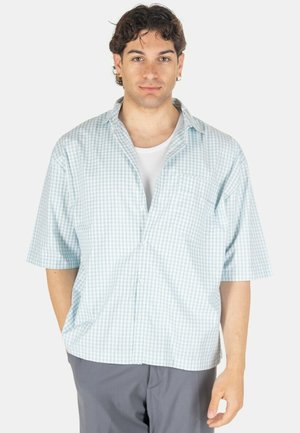 Giovane uomo che indossa una camicia a maniche corte a quadri azzurri chiari sopra una maglietta bianca e pantaloni grigi, in piedi con le mani rilassate.