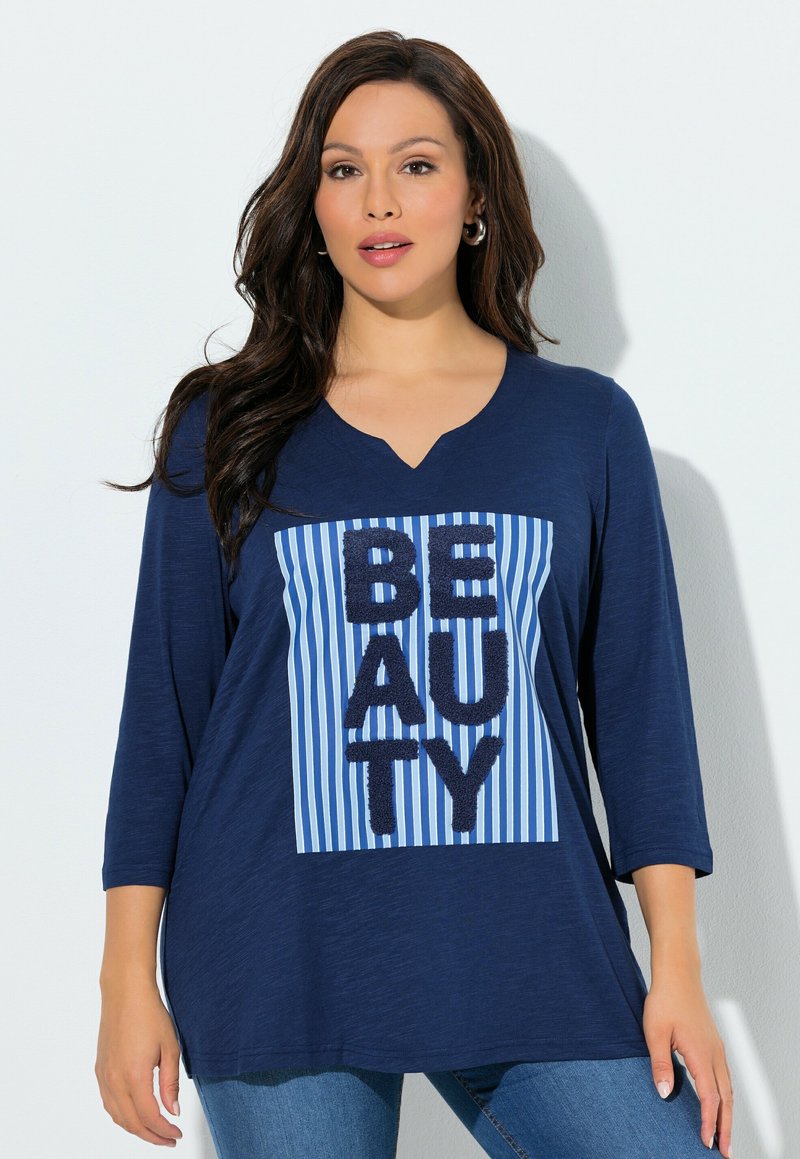 Mujer con cabello largo y oscuro, vistiendo una blusa azul marino con texto "BEAUTY" sobre un cuadrado a rayas azules y blancas, combinada con jeans azules.