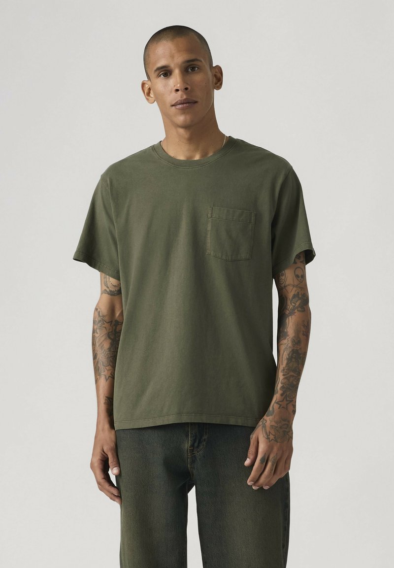 Man met tatoeages die een olijfgroen T-shirt met korte mouwen en zak en een donkere spijkerbroek draagt, staand tegen een effen lichte achtergrond.