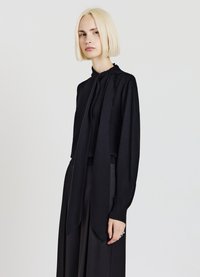 Schwarze Langarmbluse mit hohem Kragen und Schleifendetail. Weicher, glatter Stoff. Das Modell trägt sie zu hoch taillierten schwarzen Hosen.