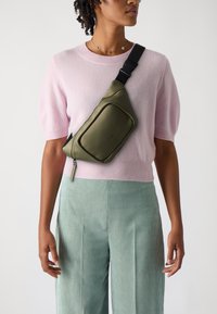 Olivgrüne Umhängetasche mit glatter Textur, geschwungenem Design und Fronttasche, getragen über einem hellrosa Pullover und gestreiften türkisfarbenen Hosen.