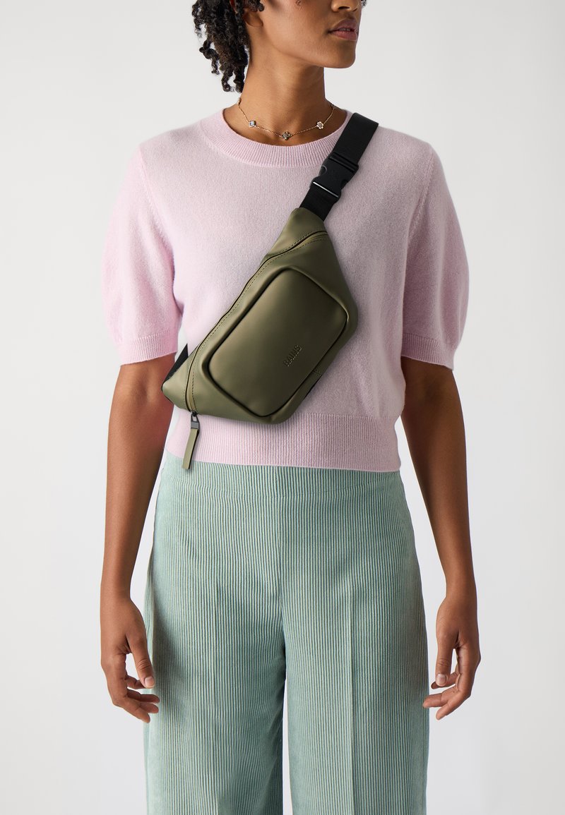 Olivgrüne Umhängetasche mit glatter Textur, geschwungenem Design und Fronttasche, getragen über einem hellrosa Pullover und gestreiften türkisfarbenen Hosen.