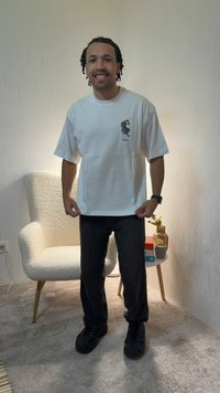 Lachend persoon in een wit T-shirt met grafisch ontwerp, zwarte broek en zwarte schoenen staat in een kamer met een witte, gestructureerde stoel en een klein rond tafeltje.