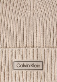 Beige knitted beanie hat featuring ribbed texture, a folded cuff, and a rectangular Calvin Klein logo tag stitched at the front.
