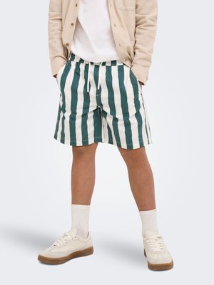 Person iført grønne og hvide vertikalt stribede shorts, beige sneakers, hvide sokker, en hvid skjorte og en beige jakke med hænderne i lommerne.