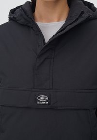 Chaqueta con capucha negra y un bolsillo frontal, hecha de un tejido ligero. Presenta un parche de logo de la marca y un cierre de cremallera cerca del cuello.