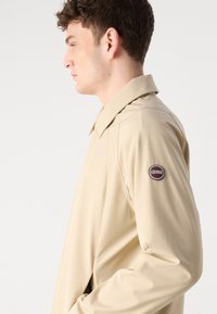 Μπεζ αδιάβροχο από λείο ύφασμα με δομημένο γιακά. Διαθέτει λογότυπο στο αριστερό μανίκι και αδιάλειπτη πλευρική λεπτομέρεια.