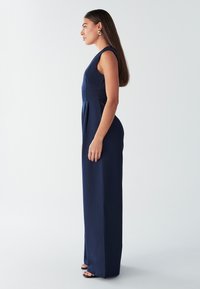 Temno modra brezrokavnik kombinezon z oprijetim zgornjim delom in širokimi hlačnicami. Gladek material; urejene gube in minimalističen slog; nošen z črnimi sandali.